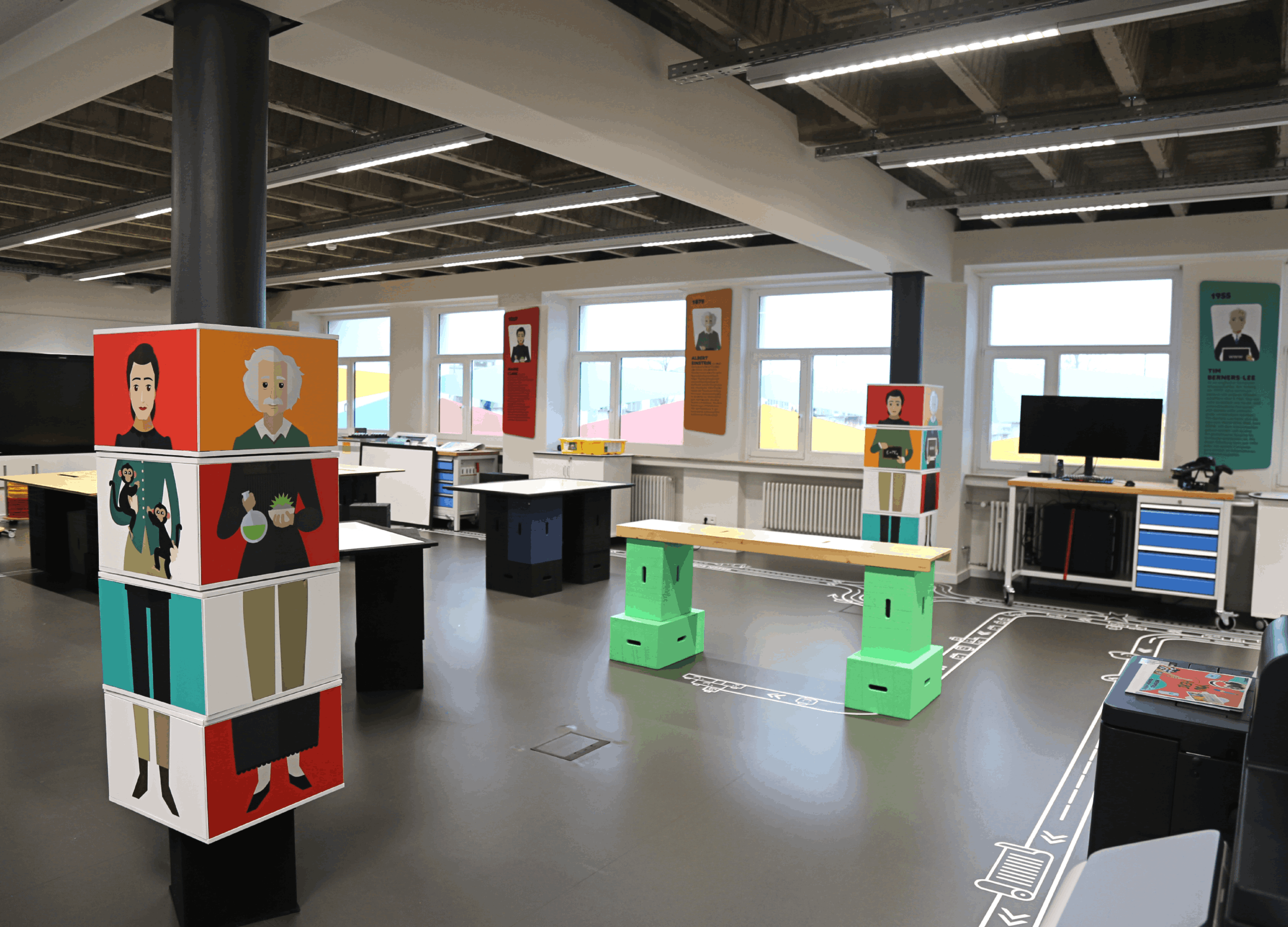 Large salle de classe moderne de type makerspace ou laboratoire d'innovation. L'espace comprend des tables de travail noires, un établi sur des tréteaux vert vif et des colonnes décorées d'illustrations colorées de scientifiques célèbres comme Albert Einstein et Marie Curie. Le sol gris est marqué de lignes blanches rappelant un circuit ou un parcours. Au fond, de grandes fenêtres apportent une lumière naturelle, et des affiches biographiques sont fixées entre elles.