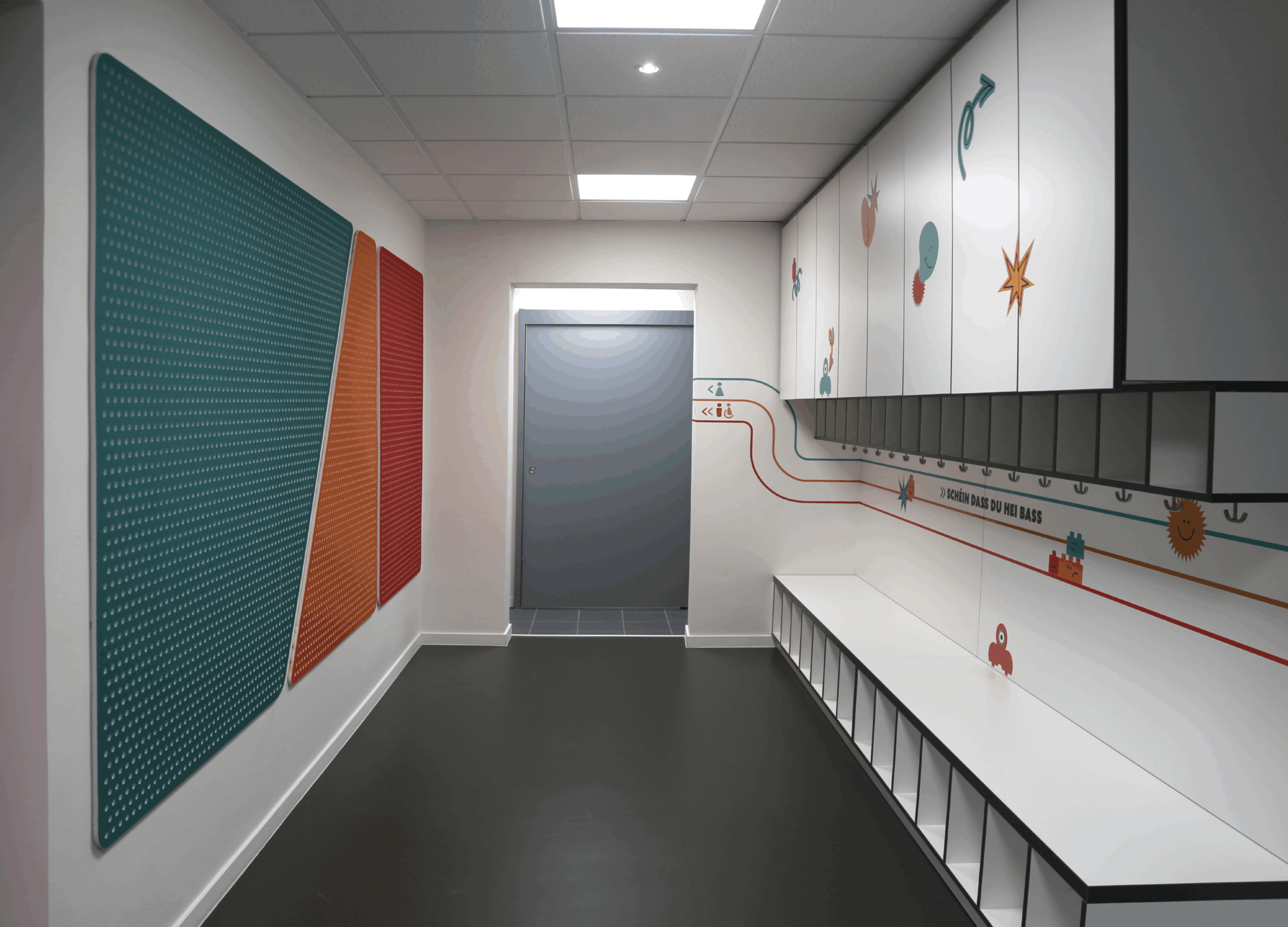 Couloir d'entrée moderne et épuré menant à un makerspace ou une structure éducative. Sur le mur de gauche, trois grands panneaux perforés colorés (bleu canard, orange et rouge) sont fixés. Le mur de droite est aménagé avec un long banc blanc surmonté de casiers et de placards de rangement blancs décorés de petits graphismes ludiques. Des lignes de signalétique colorées sur le mur mènent vers une porte grise au fond. Le sol est sombre et l'éclairage est assuré par des dalles LED au plafond.
