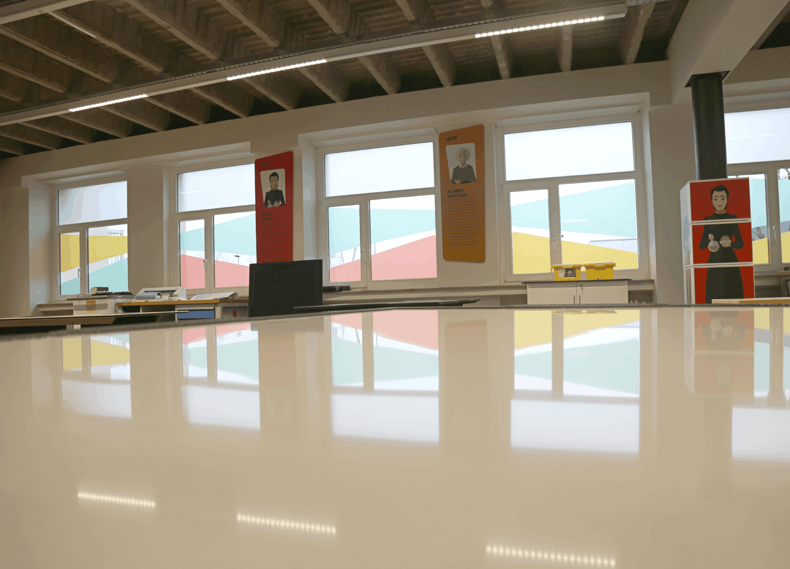 Vue en contre-plongée depuis la surface brillante d'une table blanche dans l'atelier. En arrière-plan, de grandes fenêtres avec des films colorés et des panneaux biographiques muraux (Marie Curie en rouge, Albert Einstein en orange). Le plafond révèle une structure industrielle en béton.