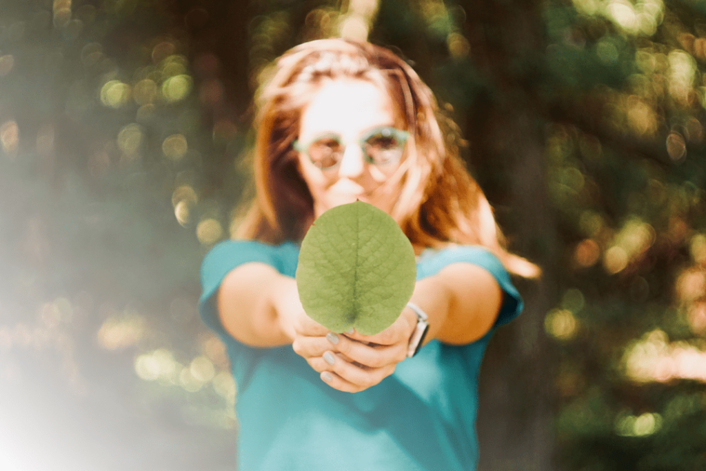 L'image montre une femme tenant une feuille verte bien en évidence vers l'objectif, avec un effet de flou sur son visage et l'arrière-plan naturel. La lumière douce et la composition centrée sur la feuille renforcent un message écologique et symbolique, mettant en avant la nature et l'engagement environnemental. L'effet de flou crée une atmosphère chaleureuse et organique.