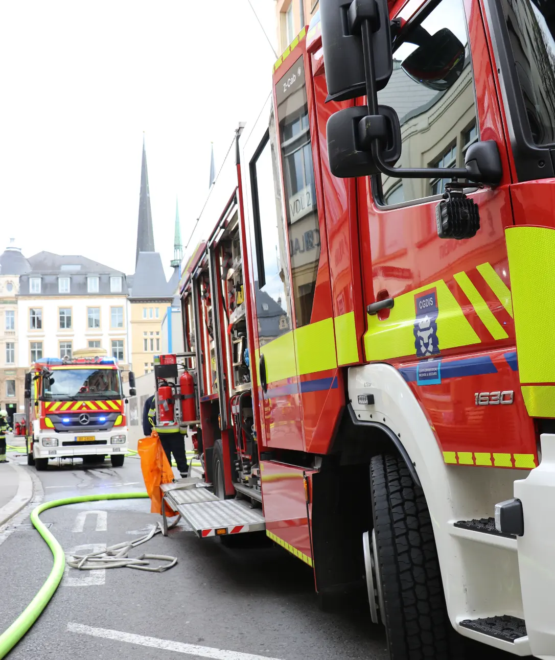 Camions de pompiers en intervention dans une rue d'une ville, probablement lors d'un exercice ou d'une situation d'urgence. Le premier plan montre un camion de pompiers rouge avec des bandes jaunes réfléchissantes, garé sur le côté de la rue avec des portes latérales ouvertes, révélant l'équipement à l'intérieur. Un tuyau d'incendie est déployé sur la chaussée, prêt à être utilisé. En arrière-plan, un autre camion de pompiers est visible, avec un bâtiment public et des flèches d'église en fond, indiquant un contexte urbain. L'image capte l'urgence et la préparation nécessaires lors des opérations de secours en milieu urbain.
