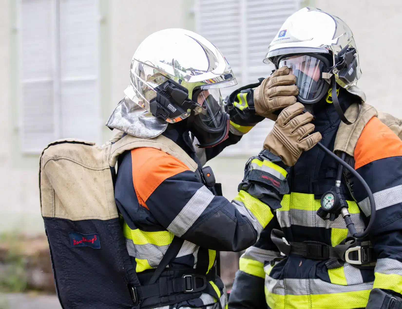 L'image montre deux pompiers en tenue complète, dont un aide l'autre à ajuster son masque. Ils portent des casques réfléchissants et des vêtements de protection adaptés aux interventions incendie.