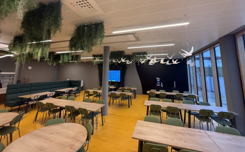 Salle de restauration rénovée avec tables en bois clair, chaises vertes, plafond végétalisé et suspensions en forme d’oiseaux blancs.