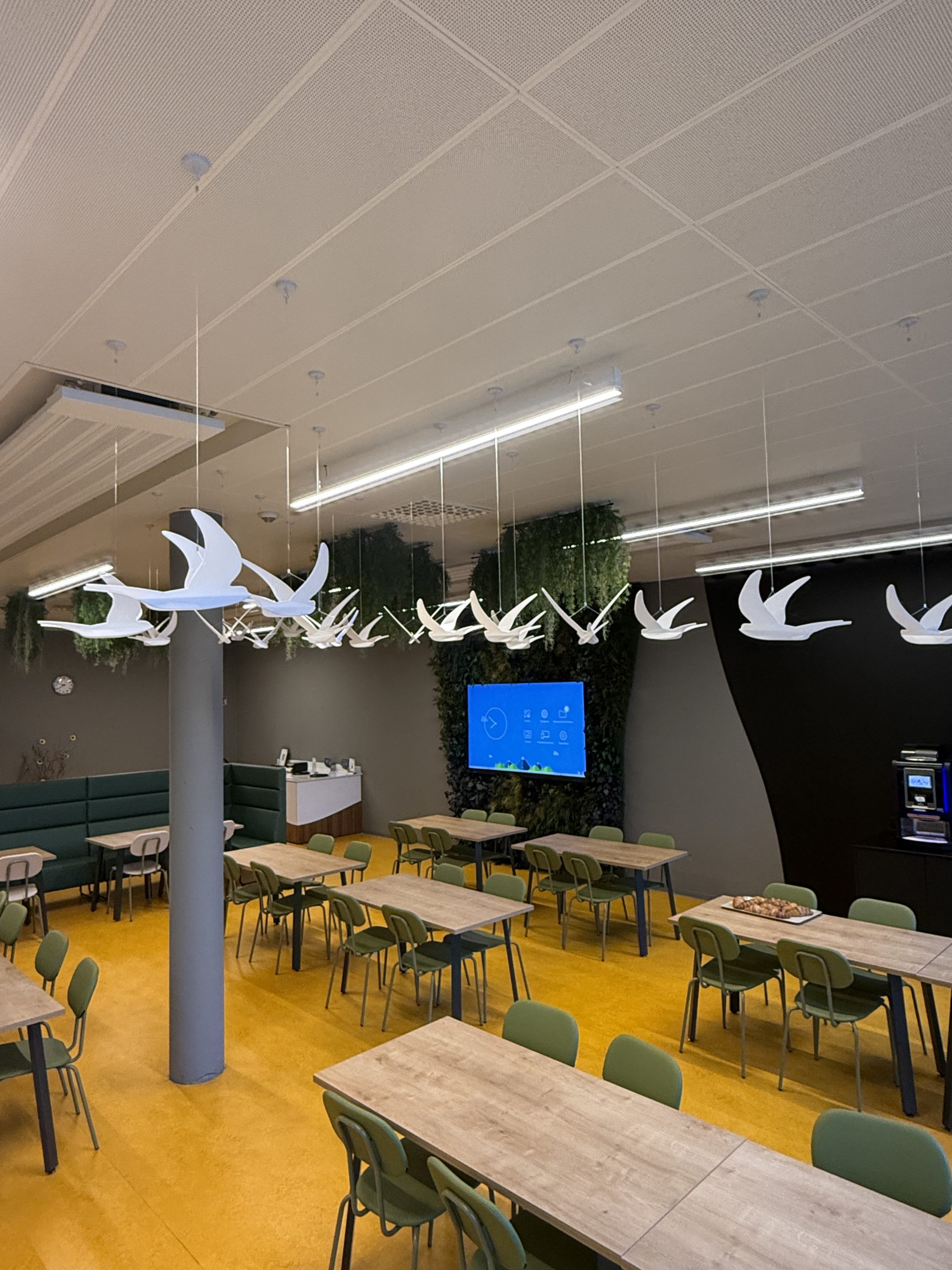 Salle collective avec téléviseurs muraux, tables alignées, plafond à bandes lumineuses.