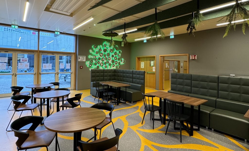 Cafétéria rénovée avec banquettes vertes, tables en bois et arbre mural lumineux vert en forme de silhouette.