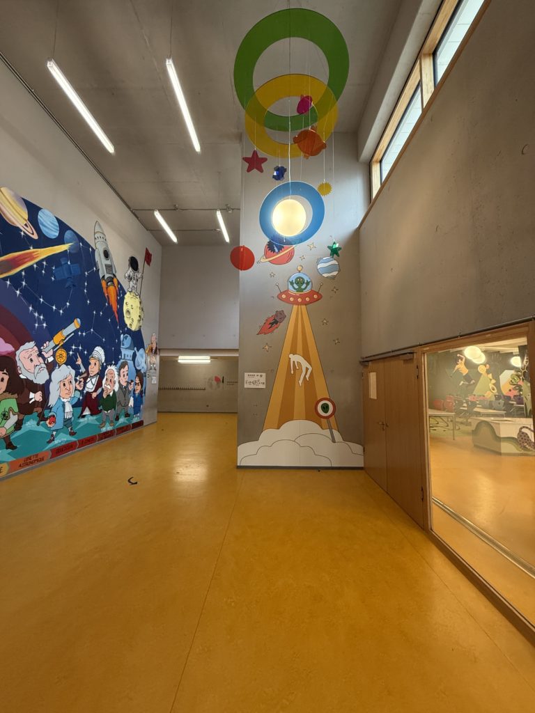 Décor mural sur le thème spatial avec fusée, planètes et personnages en apesanteur, accompagné de suspensions colorées au plafond.