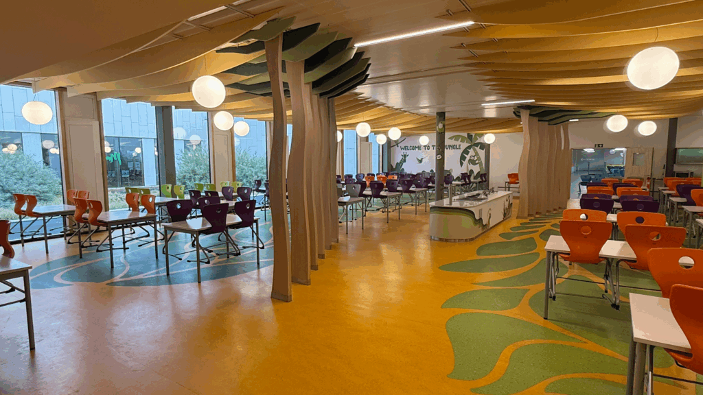 Grande cafétéria avec plafond ondulé, rangées de chaises orange et violettes, et sol aux motifs végétaux.