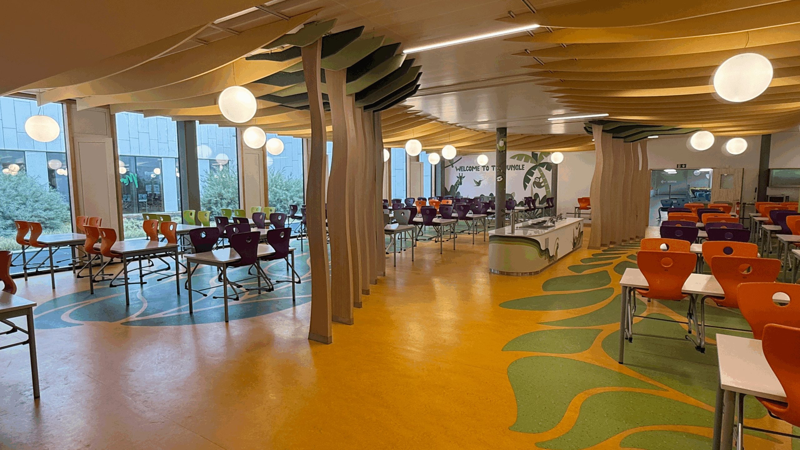 Salle collective lumineuse avec tables colorées et sol jaune, vue sur les fenêtres et colonnes design.