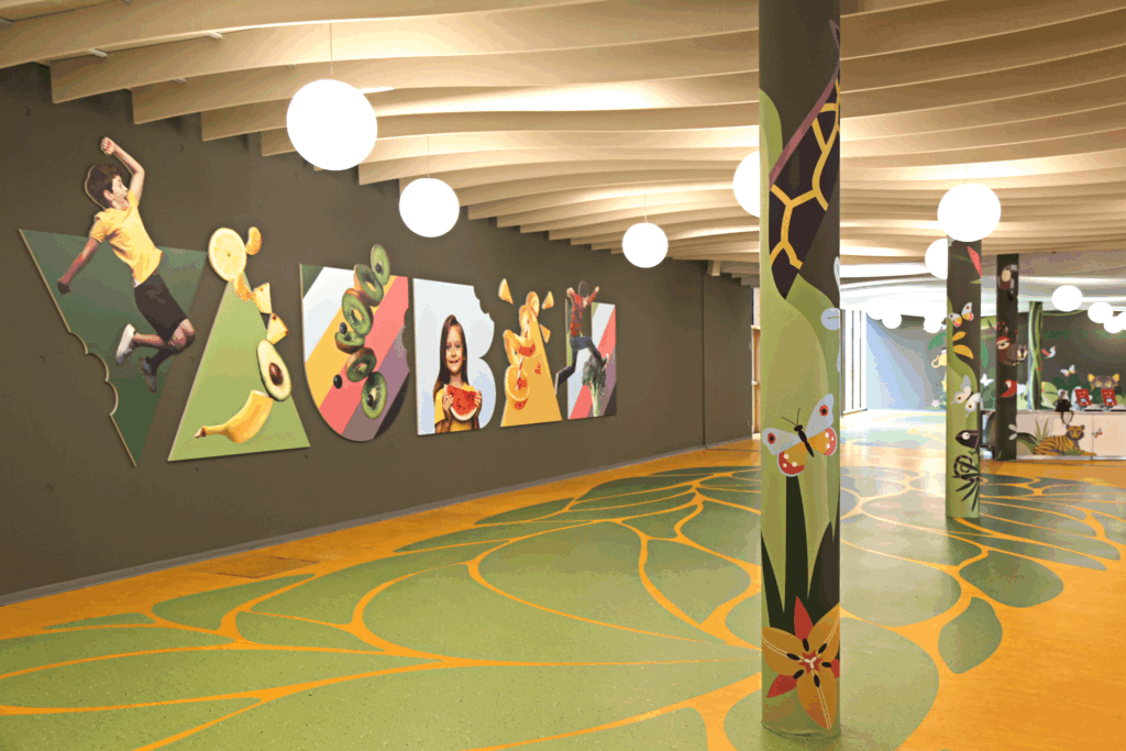 Salle de restauration décorée avec fresques murales colorées représentant fruits et personnages, colonnes peintes avec motifs nature et sol graphique vert et jaune.