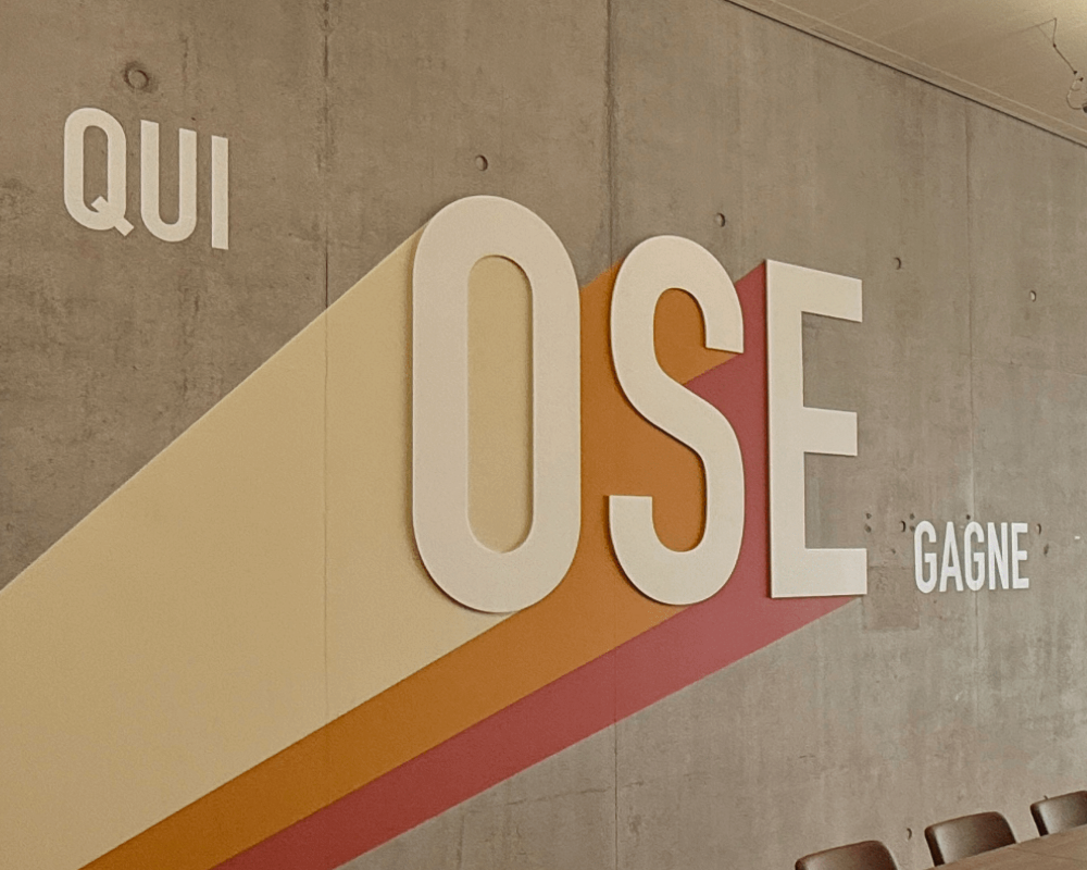 Gros plan du slogan « QUI OSE GAGNE » ; grand « OSE » en relief avec bandes obliques beige-orange-rouge ; table et chaises au premier plan