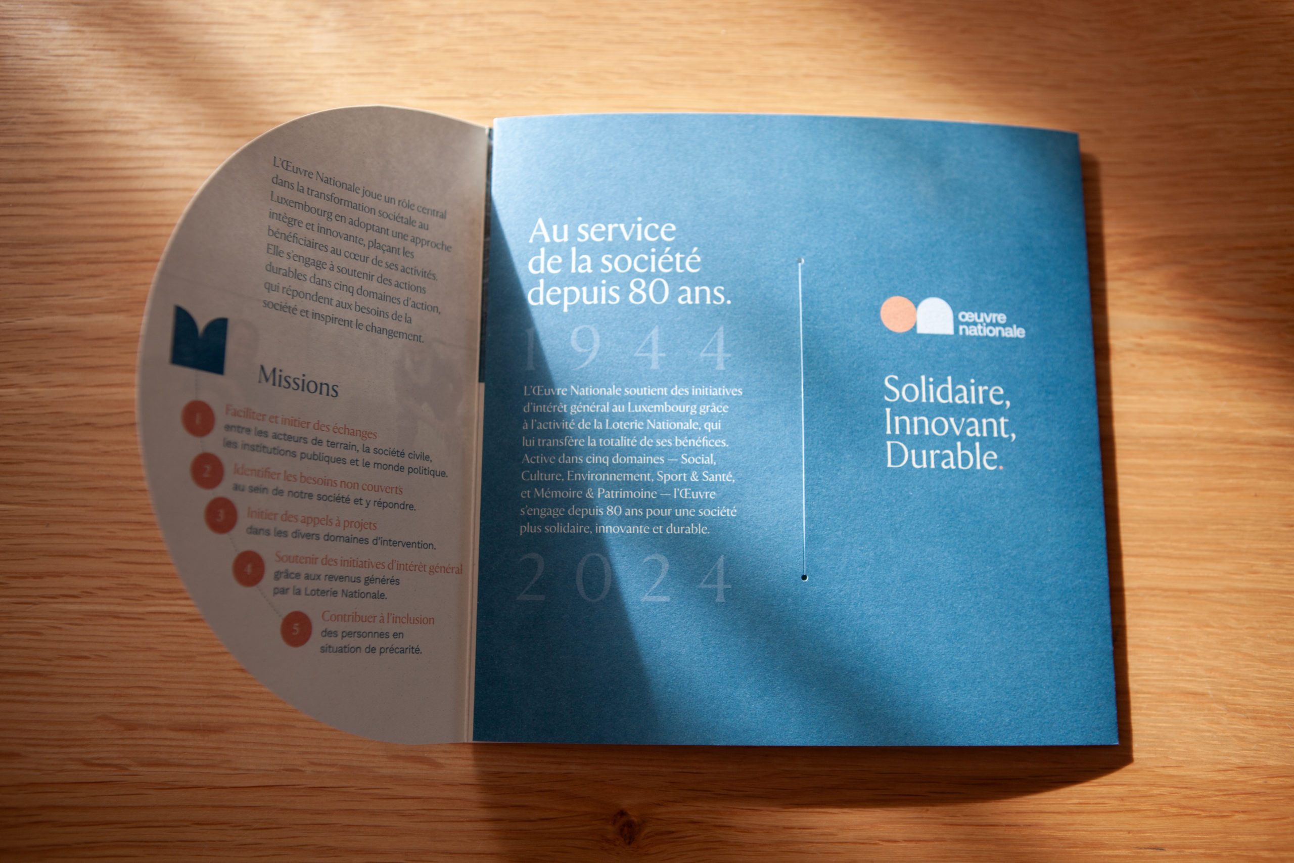 Brochure ouverte sur une table en bois avec page gauche listant les missions dune organisation et page droite bleu fonce affichant le slogan solidaire innovant durable avec une mise en page elegante sous une lumiere naturelle.