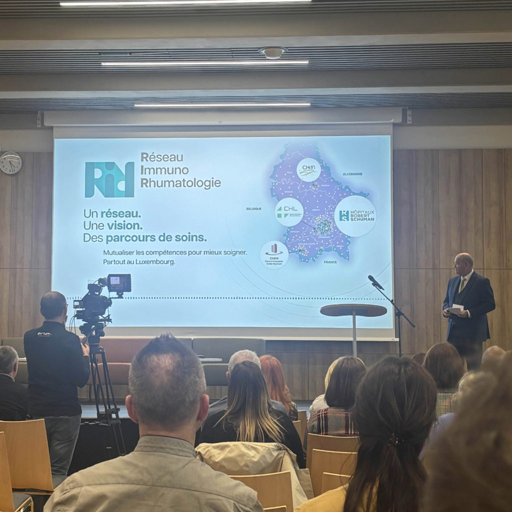 Presentation publique du RIR avec un orateur debout a droite face a une salle pleine. Un grand ecran affiche le visuel du Reseau Immuno Rhumatologie avec une carte du Luxembourg et les logos des hopitaux partenaires. Une camera filme la scene depuis la gauche.