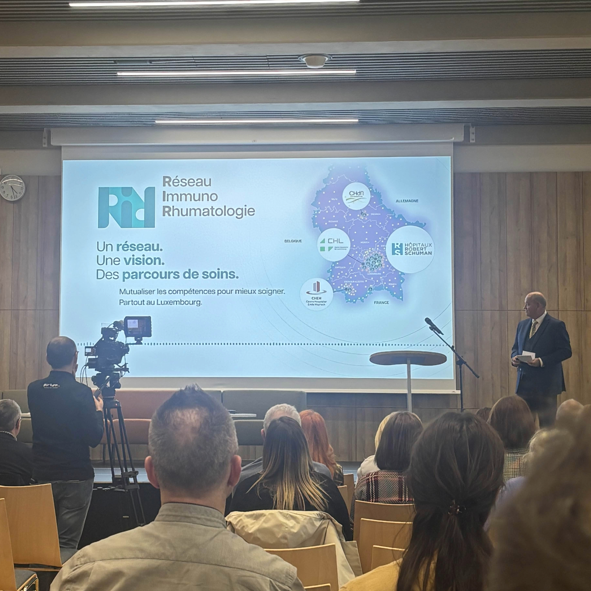 Presentation publique du RIR avec un orateur debout a droite face a une salle pleine. Un grand ecran affiche le visuel du Reseau Immuno Rhumatologie avec une carte du Luxembourg et les logos des hopitaux partenaires. Une camera filme la scene depuis la gauche.