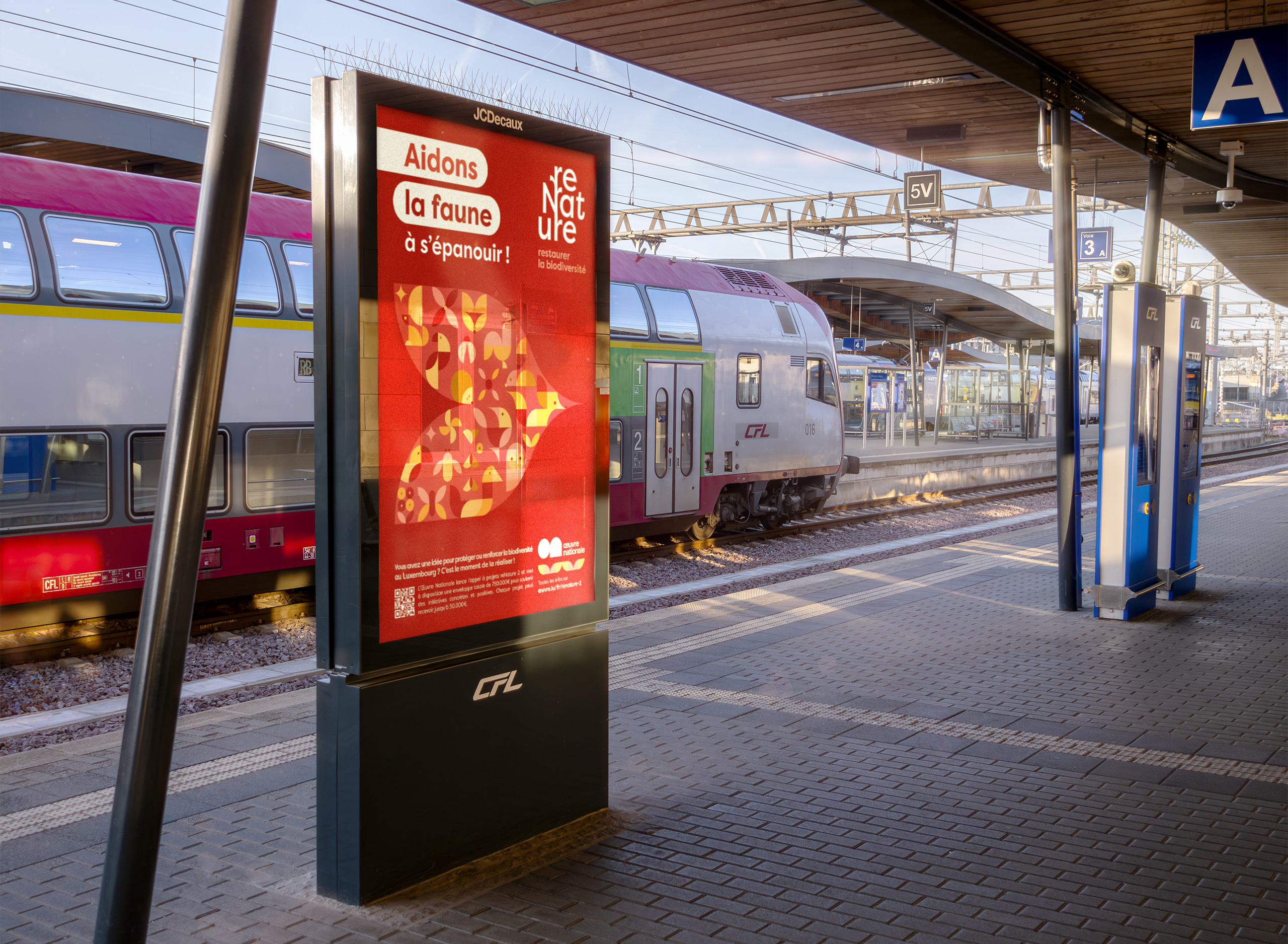 Affiche reNature sur un panneau lumineux en gare montrant un visuel rouge dedie a la faune avec un oiseau stylise. Un train CFL est a quai en arriere plan sous la lumiere du matin.