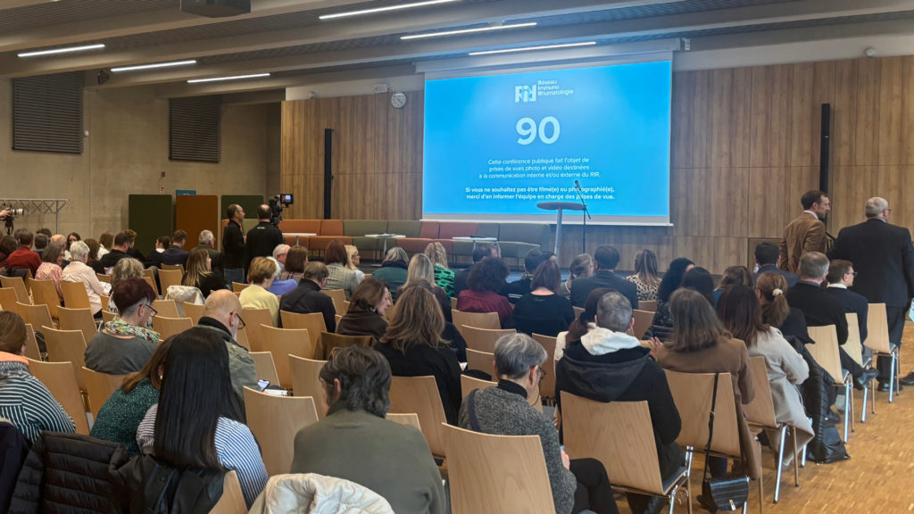 Salle de conference remplie de participants assis face a un ecran bleu affiche avec le visuel du Reseau Immuno Rhumatologie. Des personnes discutent pres de la scene et une camera filme a gauche.