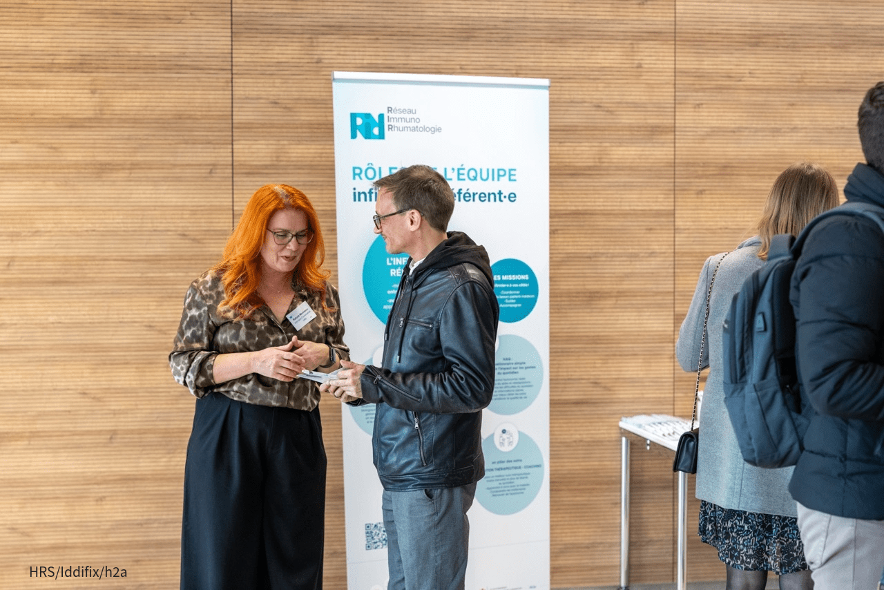 Deux personnes discutent devant un roll-up du Réseau Immuno Rhumatologie, dans un hall en bois clair. D’autres personnes sont présentes à droite, de dos.