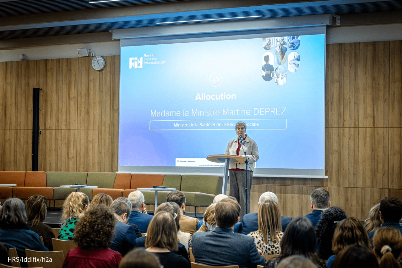 Une intervenante parle derrière un pupitre devant une salle remplie de participants. Un grand écran projette une diapositive du Réseau Immuno Rhumatologie.