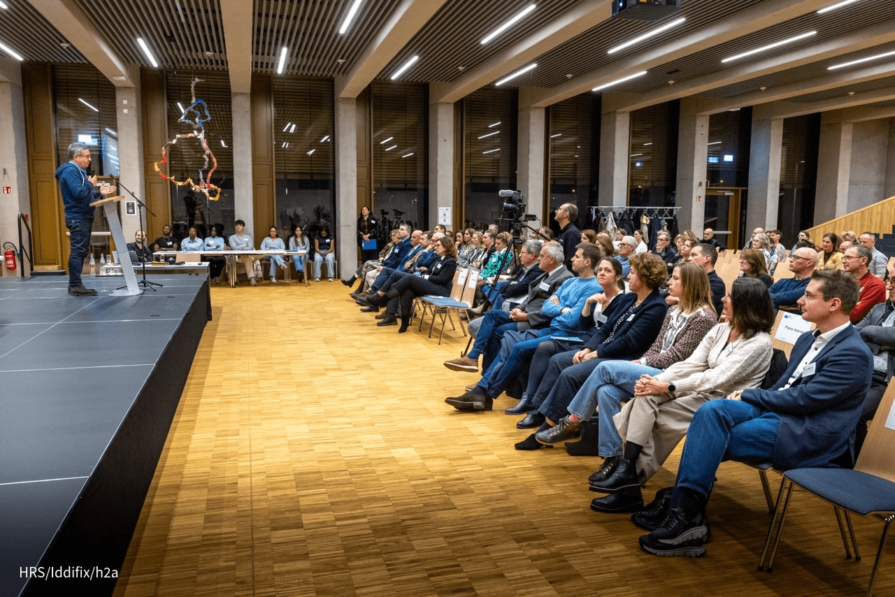 Un intervenant parle sur une scène face à un large public assis. La salle est moderne, avec plafond à lattes et grandes fenêtres.