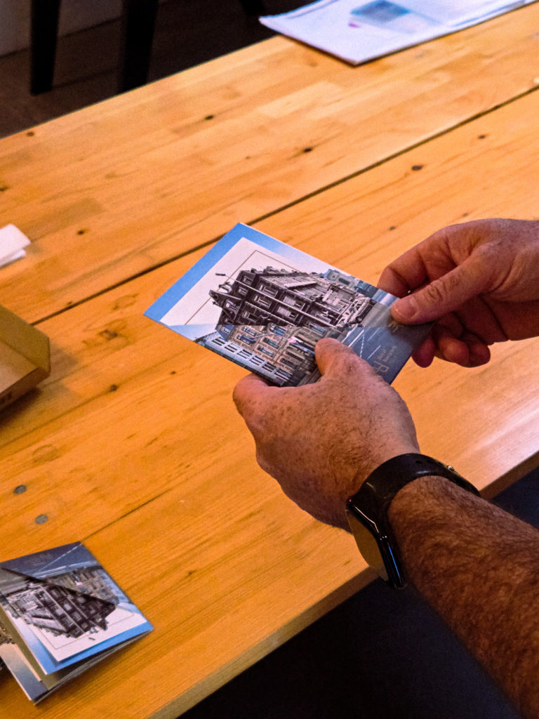 Mains tenant une brochure pliée illustrant un bâtiment historique en noir et blanc, sur une table en bois.