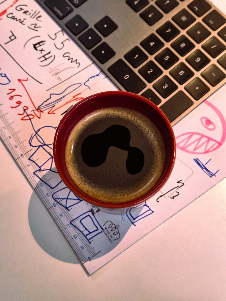 Tasse de café noir posée sur des croquis de design, des notes manuscrites, une règle et un clavier d'ordinateur.