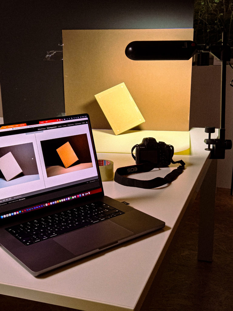 Bureau aménagé en studio photo avec un ordinateur portable affichant des images, un appareil photo et un livre éclairé par un spot.