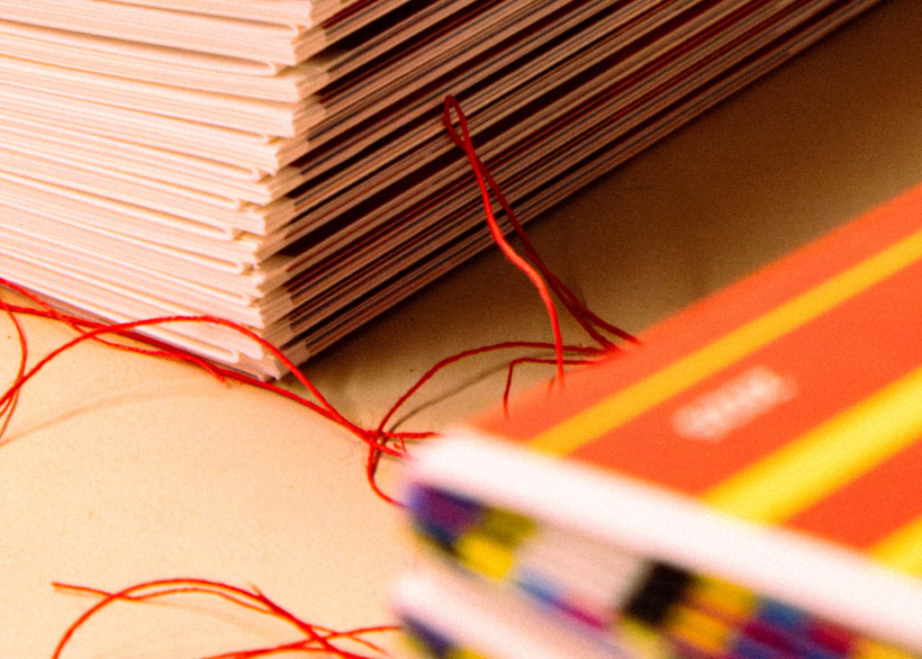 Détail technique de reliure artisanale au fil rouge sur une pile de livrets imprimés en cours de fabrication.