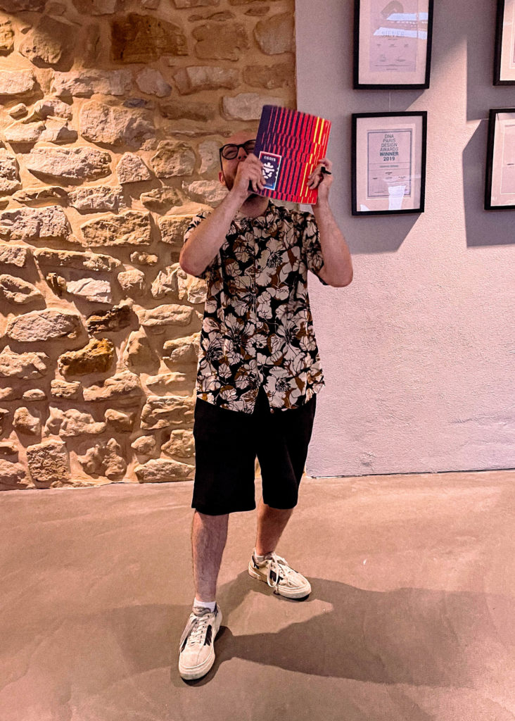 Homme en chemise à fleurs cachant son visage derrière une brochure colorée à rayures rouges et bleues, devant un mur en pierre.