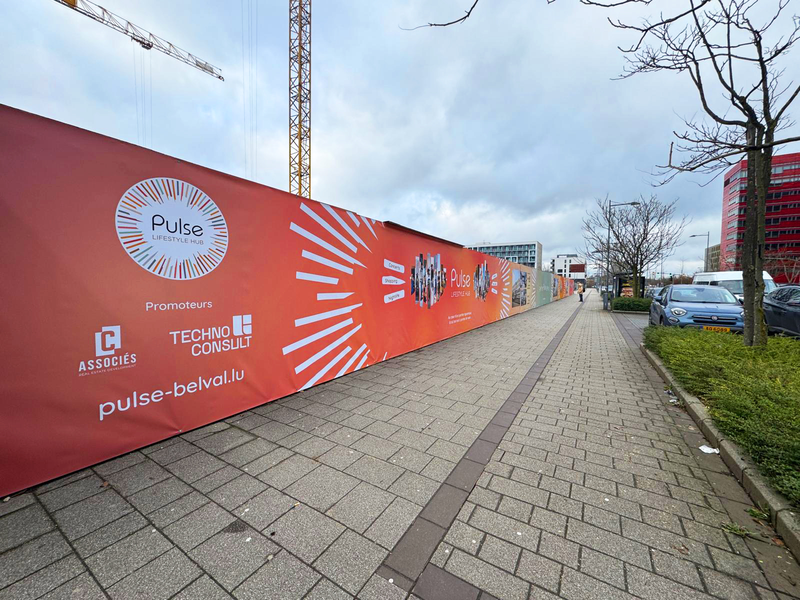 La palissade de chantier Pulse Lifestyle Hub affiche un design vif et moderne, mettant en avant le logo du projet ainsi que les noms des promoteurs, C Associés et Techno Consult. Le site internet pulse-belval.lu est clairement indiqué, renforçant la visibilité du projet. Les visuels dynamiques et les rayons graphiques apportent une énergie contemporaine, reflétant l'esprit du développement. Cette présentation donne une image professionnelle tout en valorisant l'identité du projet dans son environnement urbain.