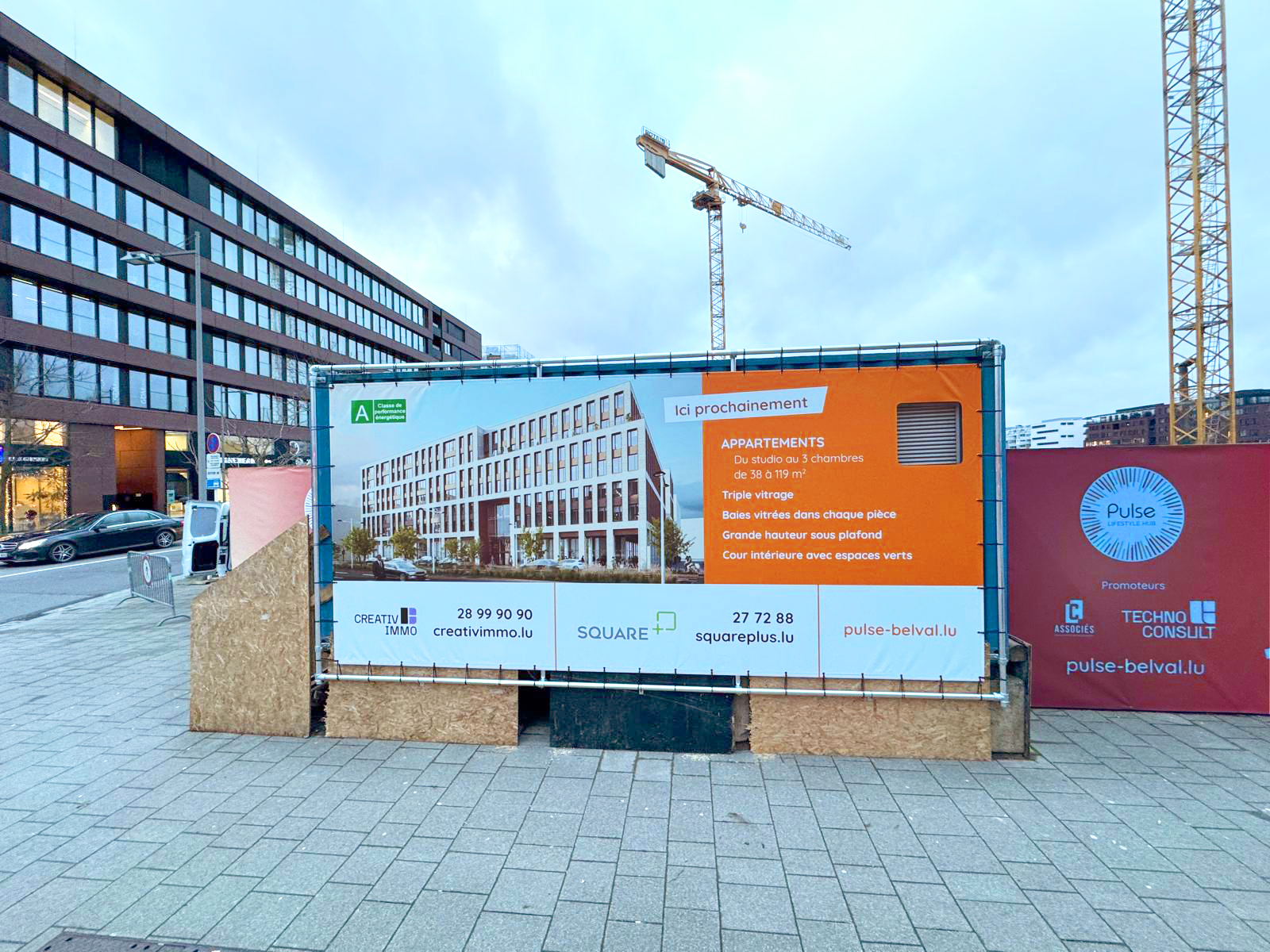 La nouvelle installation promotionnelle met en avant le projet immobilier Pulse Lifestyle Hub. Les détails incluent des appartements allant du studio aux logements de trois chambres, avec des superficies variant de 38 à 110 mètres carrés. Les caractéristiques comprennent un triple vitrage, de grandes baies vitrées pour un maximum de lumière naturelle, une hauteur sous plafond généreuse, et une cour intérieure agrémentée d'espaces verts. Les informations de contact des agences immobilières Creativ Immo et Square Plus sont clairement indiquées, tout comme le site web pulse-belval.lu, permettant aux intéressés de s'informer davantage sur le projet.