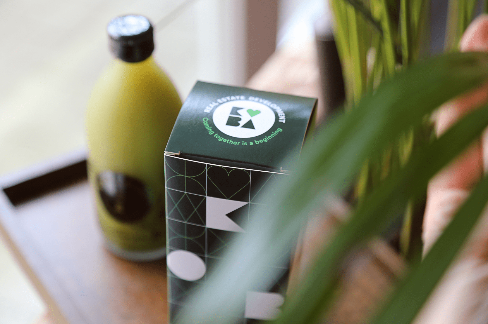 Boîte noire au design graphique minimaliste avec des formes géométriques et le logo Real Estate Development accompagné de la phrase Coming together is a beginning, posée sur une table en bois à côté d'une bouteille verte floue en arrière-plan, avec des plantes en premier plan.