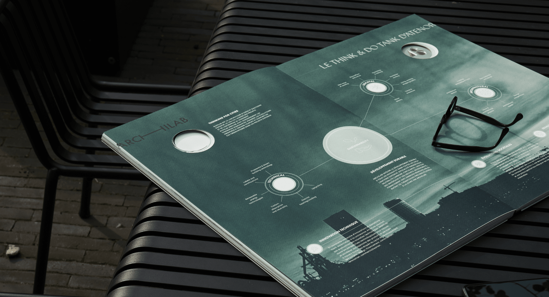 L'image montre une publication ouverte posée sur une table extérieure en métal noir, accompagnée d'une paire de lunettes. Le design graphique de la publication adopte une teinte verte sombre avec des éléments de visualisation de données et des schémas circulaires, évoquant une approche analytique et innovante. Le titre Le Think & Do Tank d'Atenor laisse entendre qu'il s'agit d'un document prospectif sur l'urbanisme ou le développement durable. L'ambiance générale, avec le fond flou et l'éclairage tamisé, renforce un sentiment de réflexion et de stratégie.