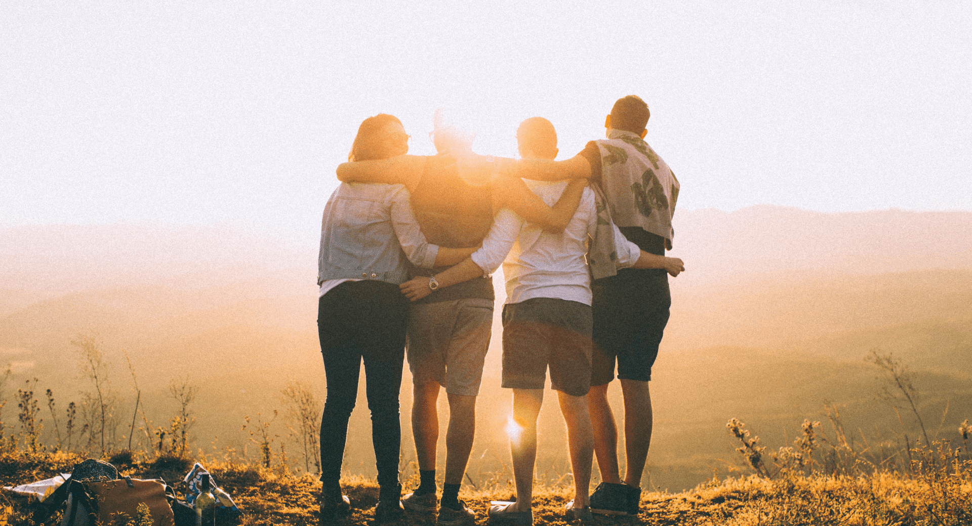 L'image montre un groupe d'amis debout sur un sommet, enlacés, face au coucher de soleil. L'éclairage chaud et doré crée une ambiance chaleureuse et nostalgique. Les silhouettes des personnes contrastent avec le paysage lumineux en arrière-plan, mettant en avant la complicité et la connexion entre elles. On perçoit une atmosphère d'aventure et de partage, renforcée par les sacs et les bouteilles posés sur le sol, suggérant un moment de détente en pleine nature.