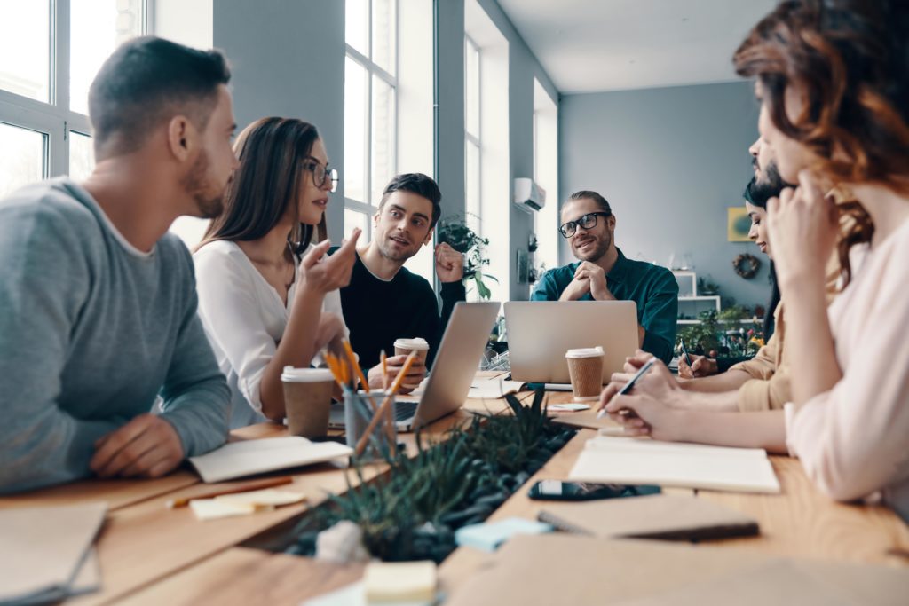 Groupe de collègues en réunion autour d’une table, discutant activement dans un bureau lumineux et moderne avec des ordinateurs portables et des carnets