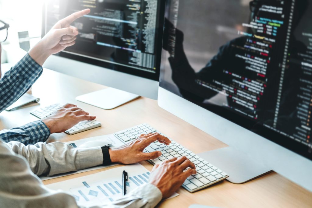 Deux développeurs travaillent côte à côte sur des ordinateurs avec du code affiché à l’écran. L’un tape au clavier pendant que l’autre pointe l’écran.