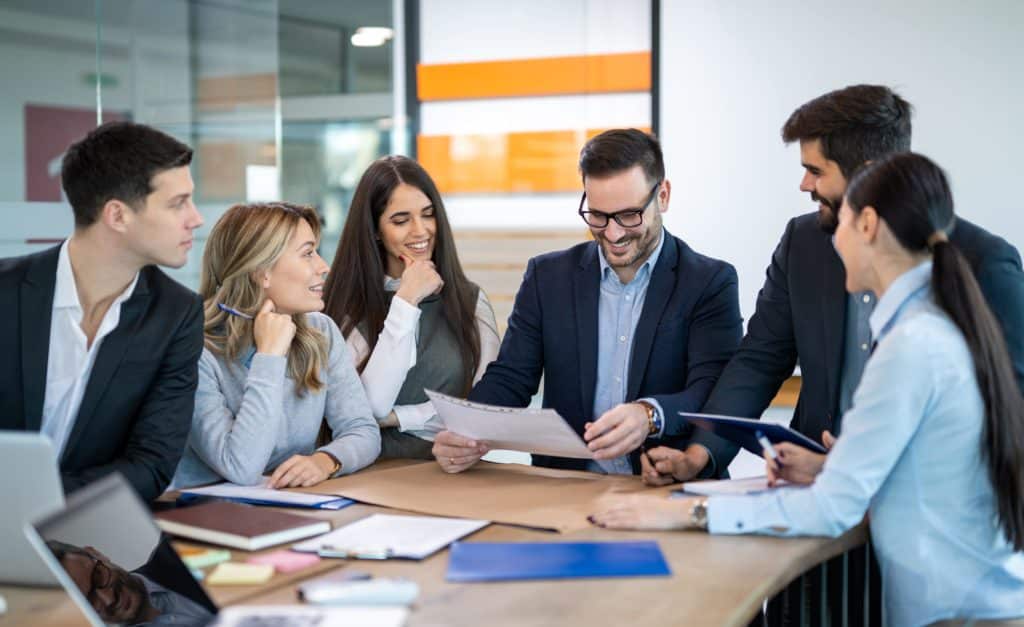 Groupe de collègues en réunion autour d’une table, discutant activement dans un bureau lumineux et moderne avec des ordinateurs portables et des carnets