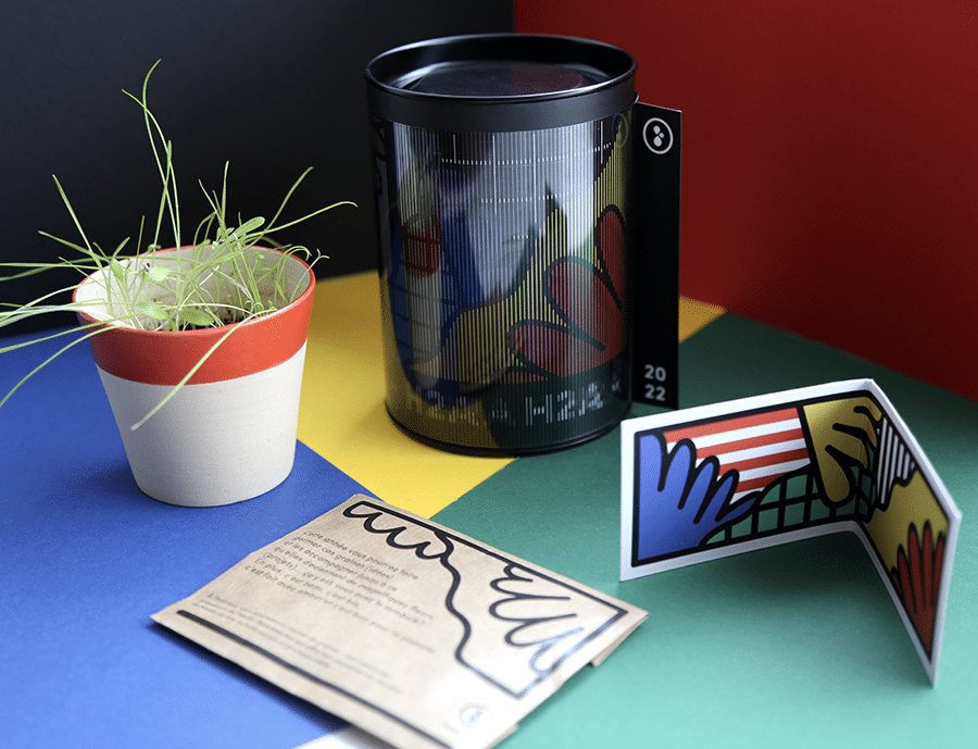 Cette image présente un ensemble d'objets colorés et graphiques sur un fond en quadrichromie. On y voit un pot en céramique bicolore avec une jeune pousse, un sachet en papier kraft avec un design minimaliste, une boîte cylindrique noire décorée avec un effet lenticulaire et la mention 2022, ainsi qu'une carte pliée illustrée avec un motif de mains et de drapeau américain. L'ensemble évoque une approche à la fois artistique et écologique, mêlant design, nature et engagement créatif.