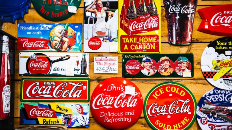 Collection d’anciennes affiches et plaques publicitaires Coca-Cola colorées, affichées sur un mur en bois, incluant des slogans rétro et des bouteilles emblématiques