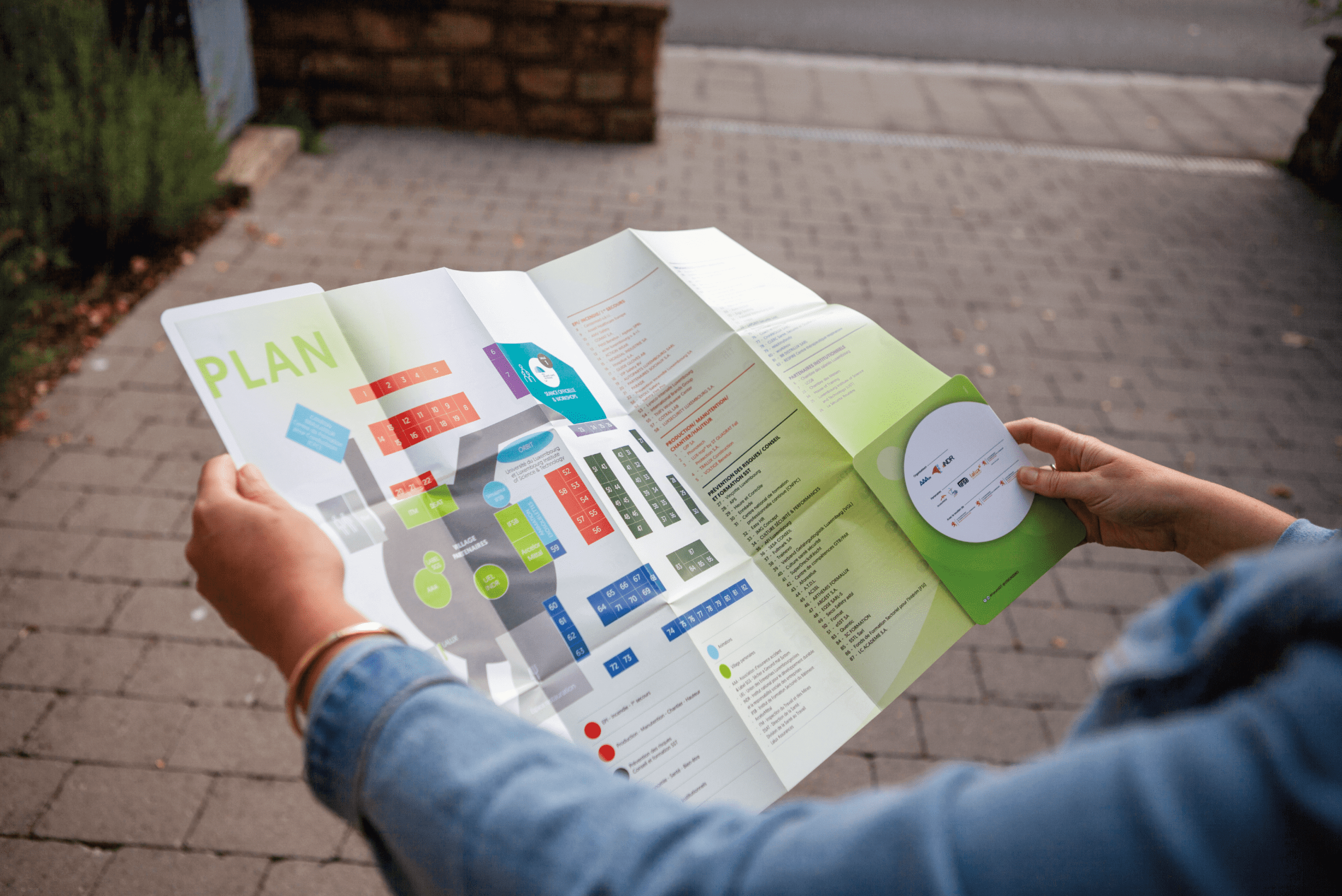 Une personne tient un plan déplié d'un événement ou d'une exposition, probablement lié à la sécurité et à la santé au travail. Le plan affiche une carte colorée avec des sections numérotées et nommées, permettant aux participants de se repérer dans l'espace de l'événement. Le titre "Plan" est bien visible en haut à gauche du document. Le plan est détaillé et inclut des informations sur les différents stands, workshops, et autres points d'intérêt. La personne semble se tenir à l'extérieur ou à l'entrée de l'événement, utilisant le plan pour naviguer ou planifier sa visite. L'ambiance est calme, avec une attention portée aux détails pour offrir une bonne orientation aux participants.