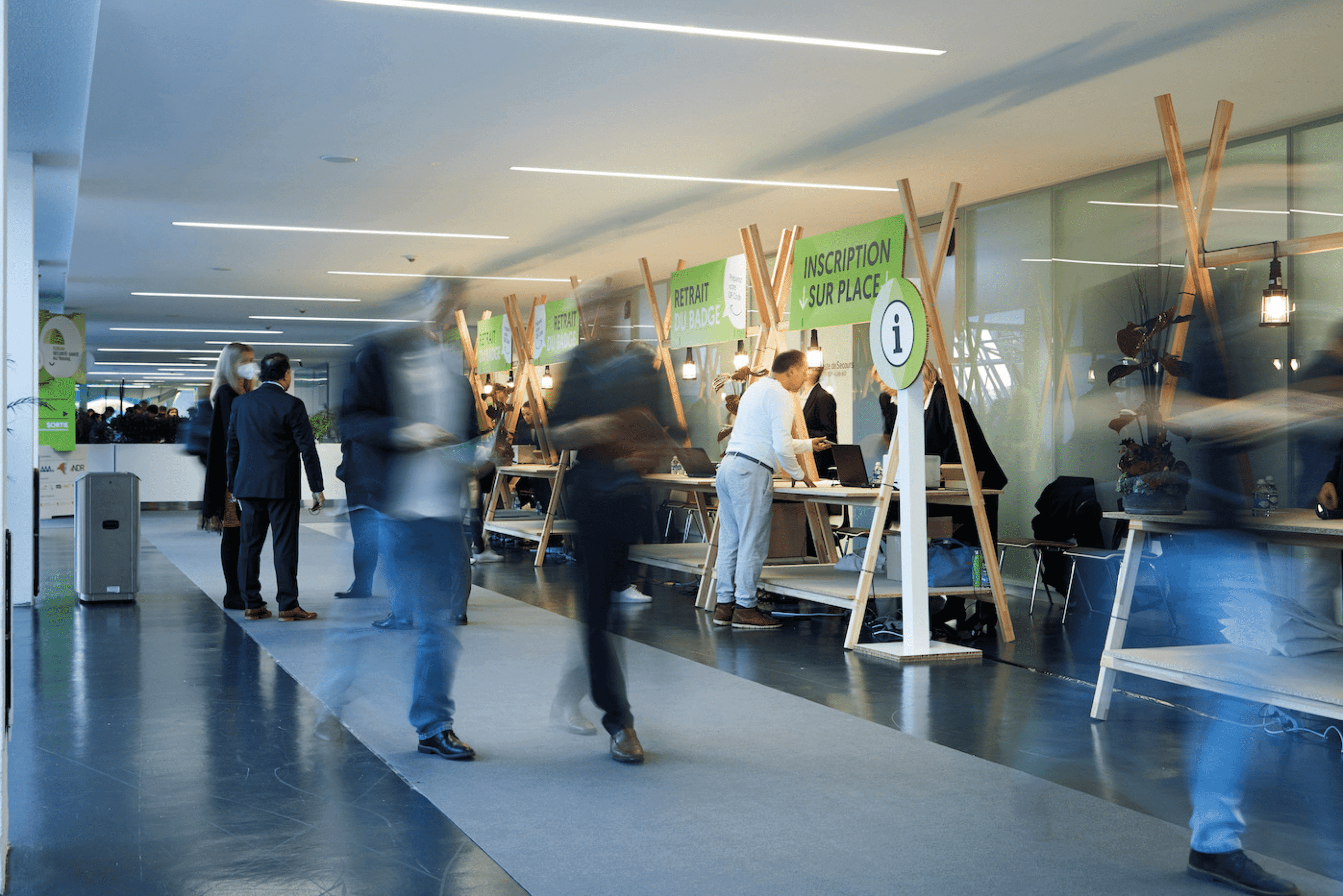Hall d'accueil d'un événement, probablement une conférence ou un forum, avec des participants en mouvement se dirigeant vers des stands d'inscription et d'information. Les comptoirs sont alignés le long du mur de droite, avec des panneaux verts indiquant des services tels que l'inscription sur place, le retrait de badge, et des informations générales. L'atmosphère est animée, avec des personnes interagissant avec le personnel ou se déplaçant dans l'espace. Le design des stands est moderne, avec des structures en bois et un éclairage chaleureux, offrant un accueil organisé et efficace pour les participants.