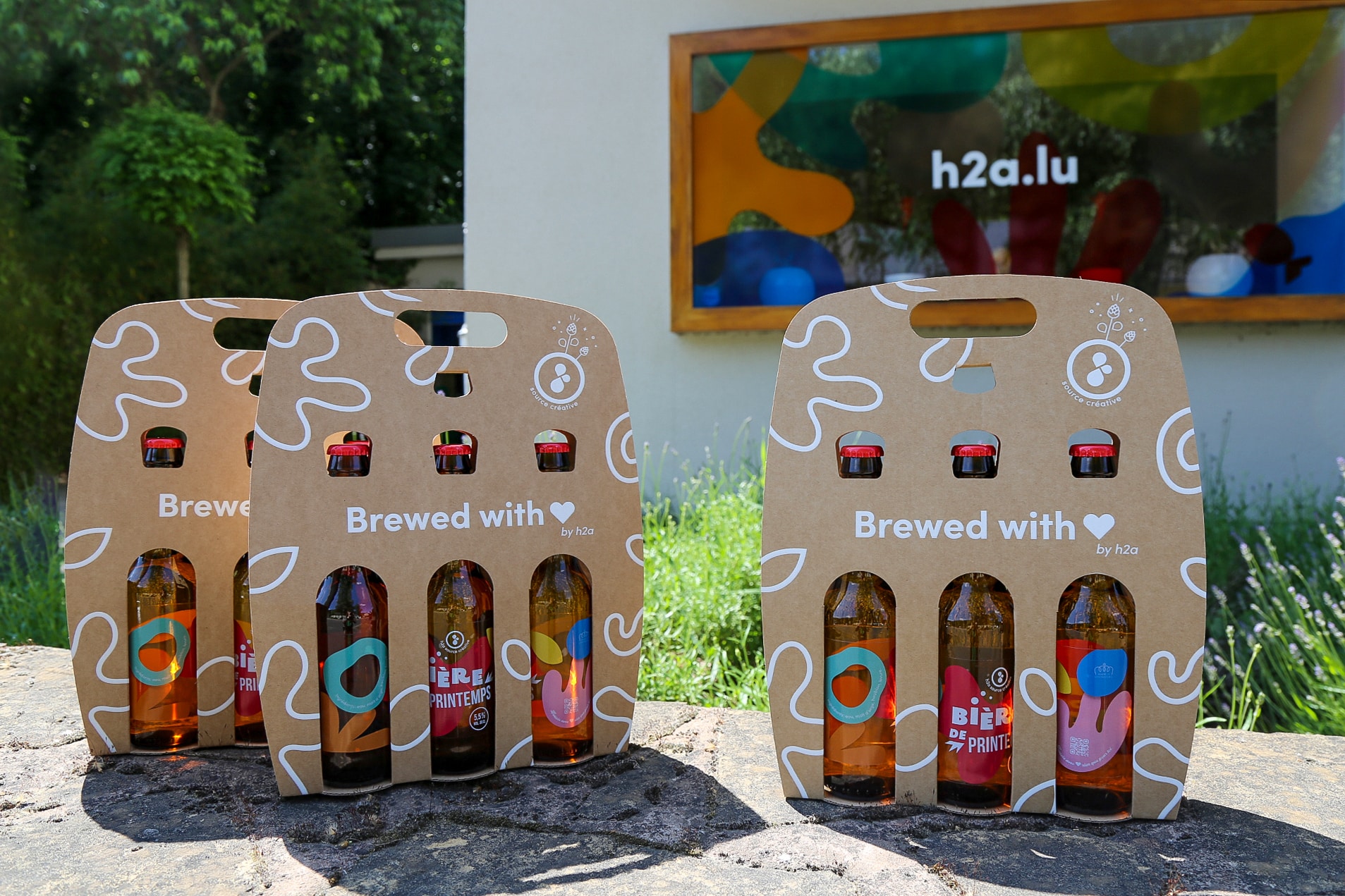 Trois packs de six bouteilles de bière artisanale avec l’inscription Brewed with ❤️ by h2a, posés sur une pierre, avec en arrière-plan un panneau coloré affichant l'URL h2a.lu