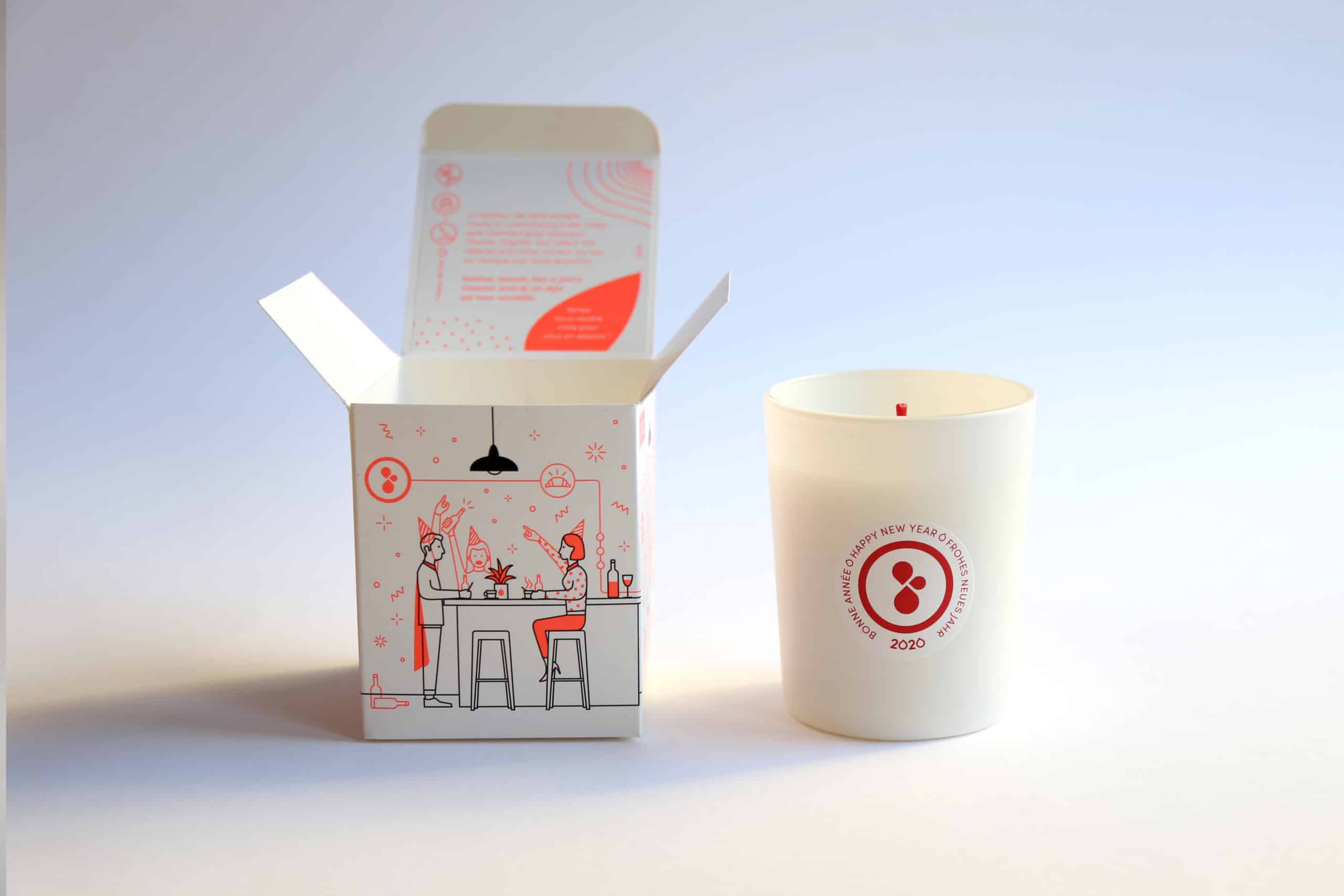 Bougie blanche avec l'inscription Bonne Année 2020 et une boîte ouverte ornée d'une illustration festive de personnes célébrant autour d'une table, dans un style minimaliste rouge et noir sur fond clair.