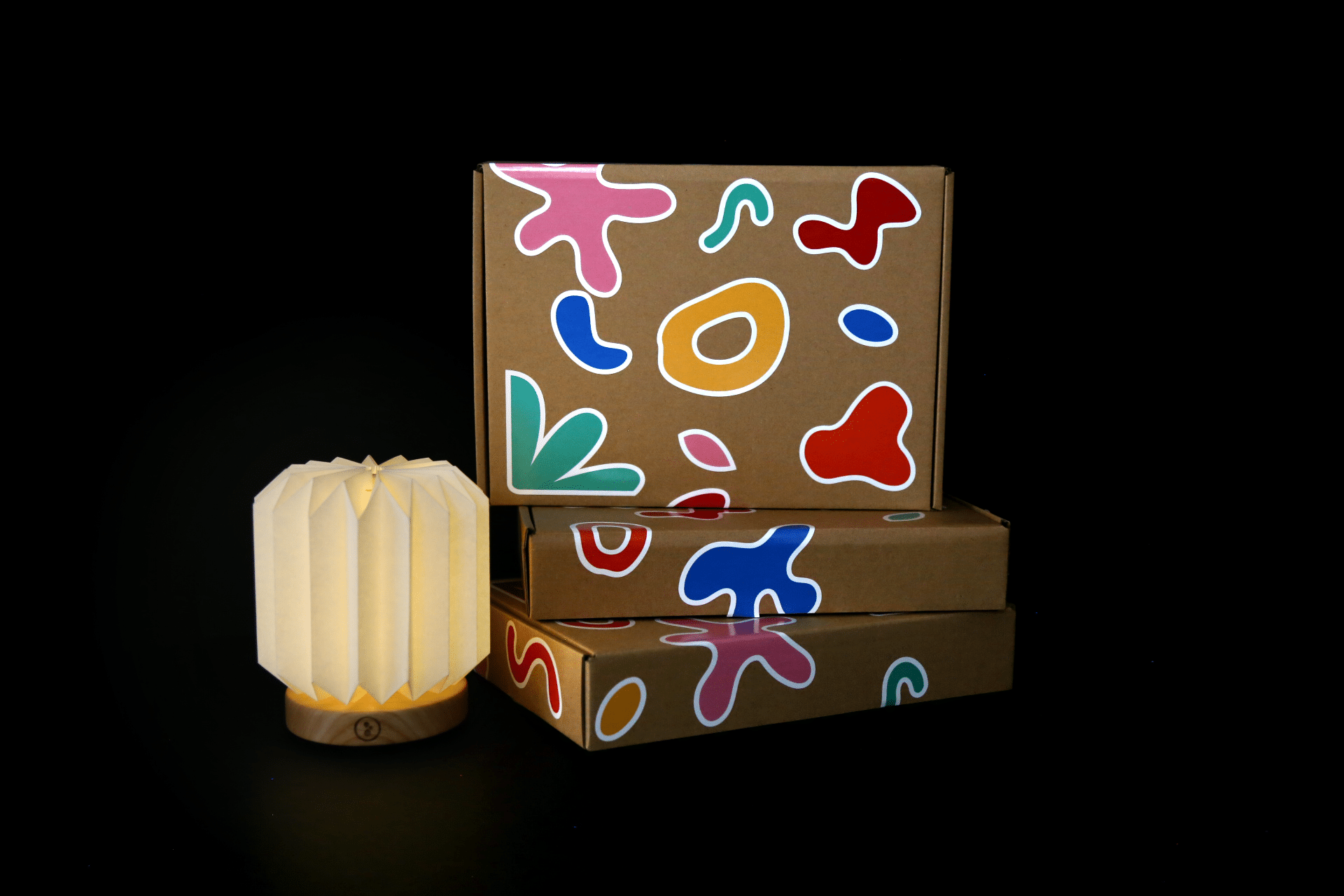 Trois boîtes en carton ornées de motifs colorés abstraits, empilées à côté d'une lampe plissée blanche avec une base en bois, sur un fond noir avec une lampe à côté