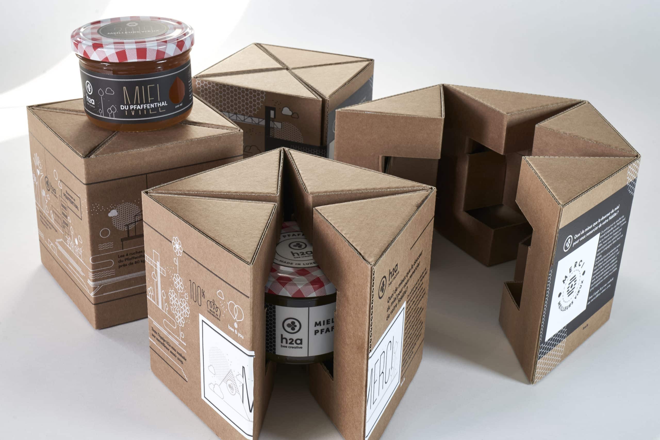 Plusieurs emballages en carton brun, chacun conçu pour contenir un pot de miel du Pfaffenthal, avec des motifs et messages imprimés. Un pot de miel à couvercle à carreaux rouges est placé sur l'un des emballages, mettant en valeur le design protecteur et esthétique des boîtes.