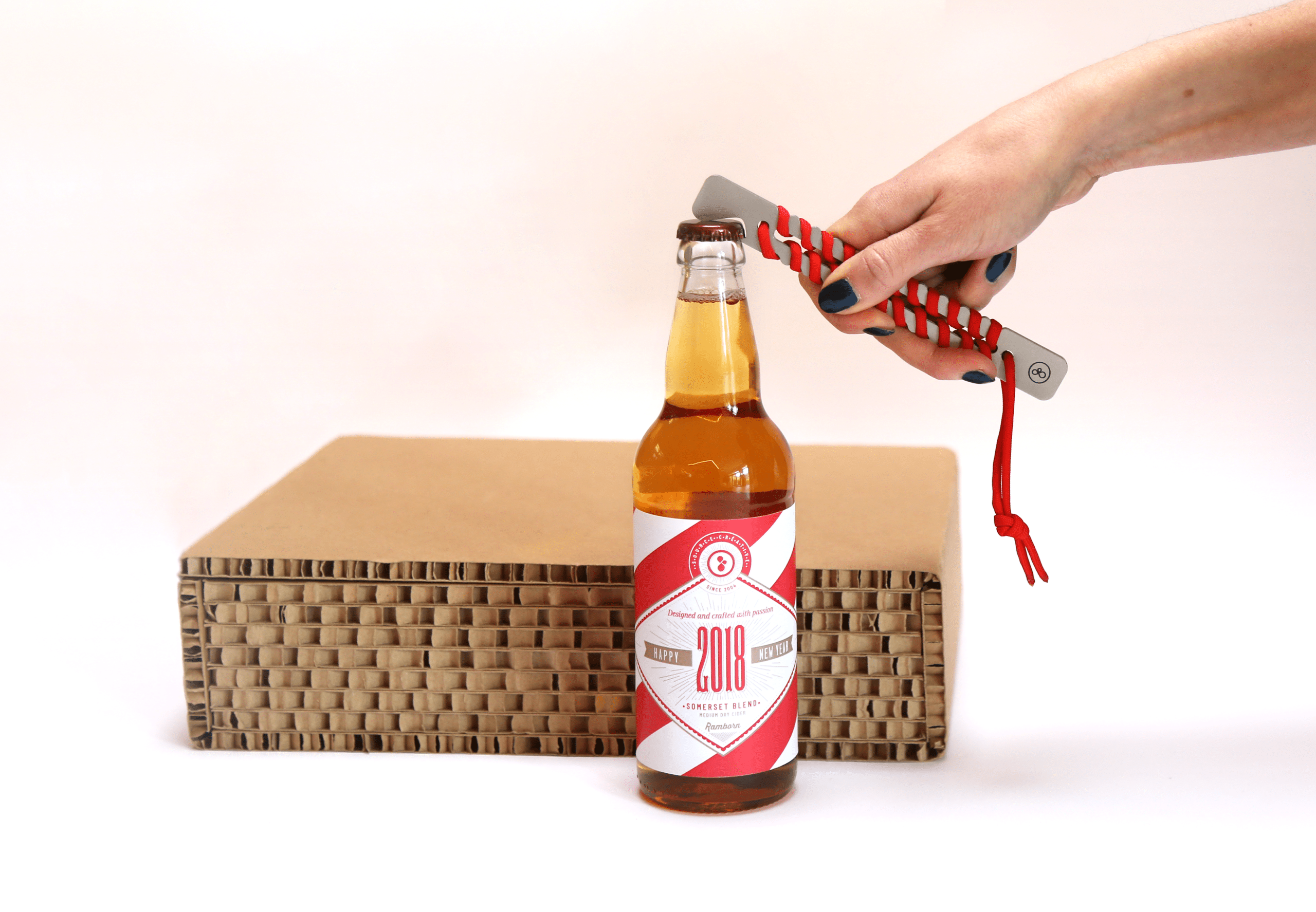 Main ouvrant une bouteille de bière millésimée 2018 avec un décapsuleur au manche tressé rouge et gris, posée devant une boîte en carton ondulé, dans un style élégant et épuré.