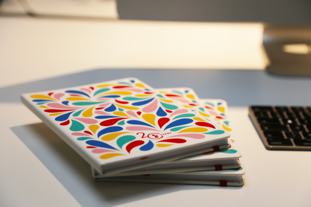 Trois carnets empilés sur une surface blanche. Les couvertures des carnets présentent un motif coloré et dynamique de gouttes en forme de larmes dans des teintes de rouge, bleu, jaune, rose et vert, avec l'inscription "20 YEARS" en bas à gauche. La lumière douce éclaire la surface, mettant en valeur les couleurs vives et les détails texturés des motifs. En arrière-plan, un clavier noir et une partie d'un moniteur d'ordinateur sont visibles, ajoutant un contexte de bureau à la scène.
