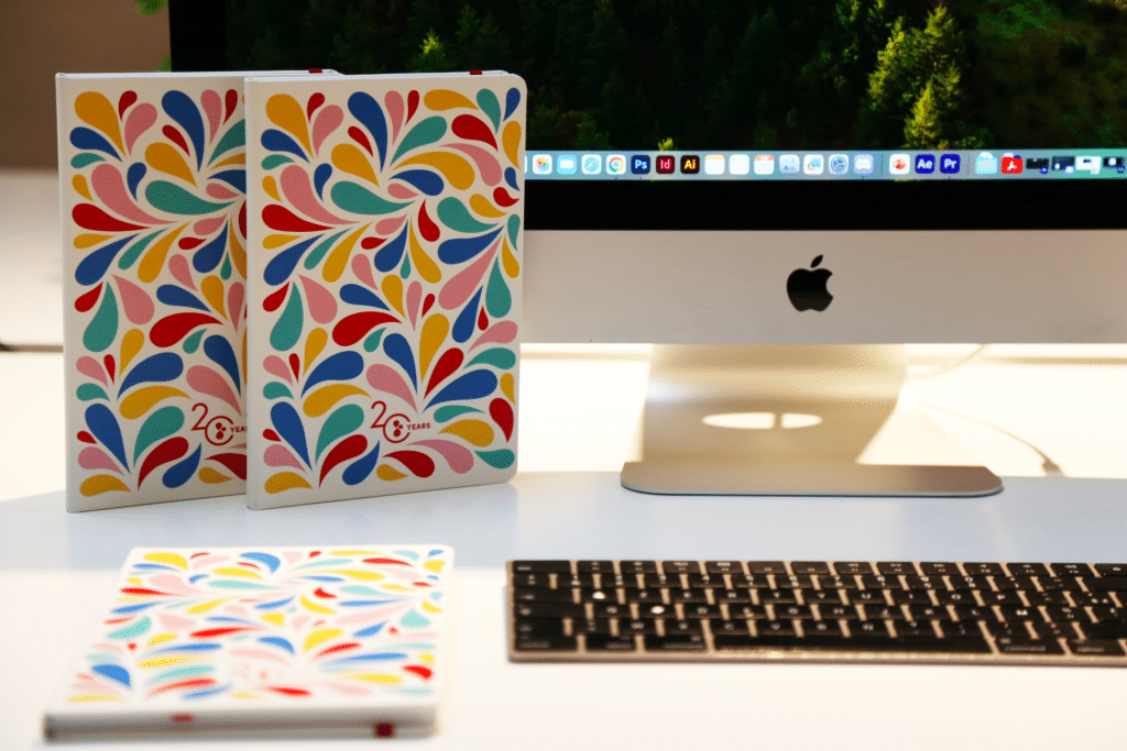 Trois carnets à couverture colorée avec des motifs de gouttes multicolores sont disposés sur un bureau blanc. Deux des carnets sont debout à côté d'un écran d'ordinateur iMac affichant une rangée d'icônes d'applications de design en haut de l'écran. Le troisième carnet est posé à plat sur le bureau devant un clavier d'ordinateur noir. Chaque carnet comporte l'inscription "20 YEARS" en rouge sur la couverture. Le bureau est bien éclairé, créant une ambiance de travail moderne et créative.