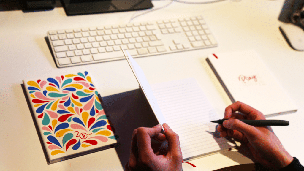 Une main écrit dans un carnet ouvert avec des lignes à l'intérieur, à l'aide d'un stylo noir. À gauche du carnet ouvert, un autre carnet avec une couverture colorée ornée de motifs de gouttes multicolores et l'inscription "20 YEARS" est posé sur la table. À droite, un troisième carnet blanc avec l'inscription "Play HARD" en rouge est partiellement visible. En arrière-plan, un clavier blanc et un écran d'ordinateur sont présents, ajoutant un contexte de bureau à la scène. La surface de la table est blanche, créant un environnement lumineux et ordonné.
