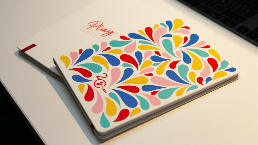 Deux carnets empilés sur une surface blanche. Le carnet du dessus présente un motif coloré et vibrant de gouttes en forme de larmes dans des teintes de rouge, bleu, jaune, rose et vert, avec l'inscription "20 YEARS" en bas à gauche. Le carnet en dessous, partiellement visible, porte l'inscription "Play Hard" en rouge. La lumière douce met en valeur les couleurs éclatantes et les détails texturés des couvertures des carnets, tandis qu'un ordinateur portable noir est visible en arrière-plan.