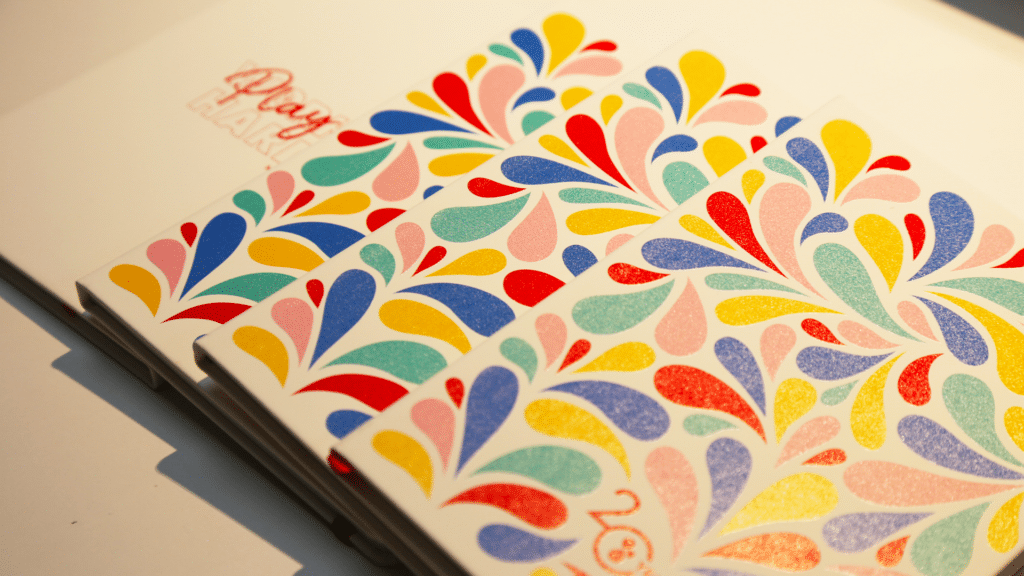 Une vue rapprochée de carnets empilés sur une surface blanche. Le carnet à l'avant présente un motif coloré et vibrant de gouttes en forme de larmes dans des teintes de rouge, bleu, jaune, rose, et vert. Le carnet en arrière-plan, partiellement visible, porte l'inscription "Play Hard" en rouge. La lumière douce met en valeur les couleurs éclatantes et les détails texturés des couvertures des carnets.
