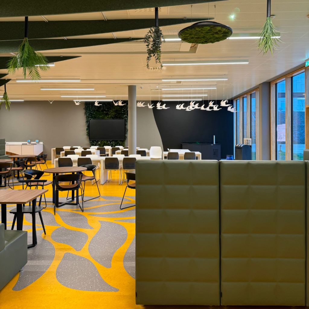 Espace de cafétéria avec banquettes vertes, tables en bois et sol graphique jaune et gris décoratif.